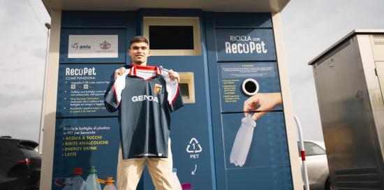 Il giocatore del Genoa Johan Vásquez davanti ad un ecocompattatore, con in mano la maglietta eco-friendly rossoblù
