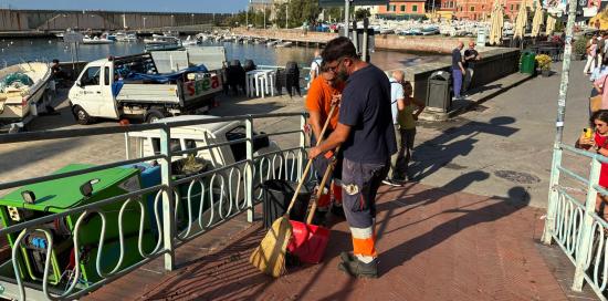 Due peratori Amiu puliscono con scopa e paletta i primi metri di passeggiata lato porticciolo Due peratori Amiu puliscono con scopa e paletta i primi metri di passeggiata lato porticciolo