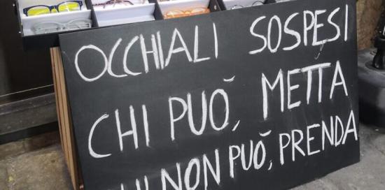 Scaffale con occhiali in esposizione e la scritta "Occhiali sospesi: chi può metta, chi non può prenda"