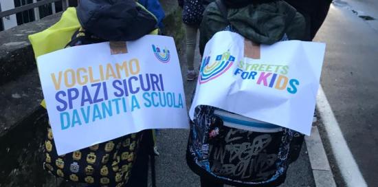 Due bimbi inquadrati di spalle durante il pedibus reggono alcuni cartelli che chiedono spazi sicuri davanti a scuola