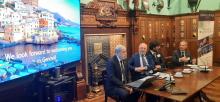 Il sindaco Bucci accanto a uno schermo dove appare una scritta che invita gli astanti a venire a visitare Genova