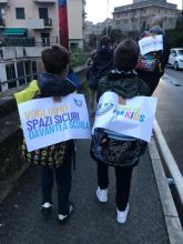 Due bimbi inquadrati di spalle durante il pedibus reggono alcuni cartelli che chiedono spazi sicuri davanti a scuola